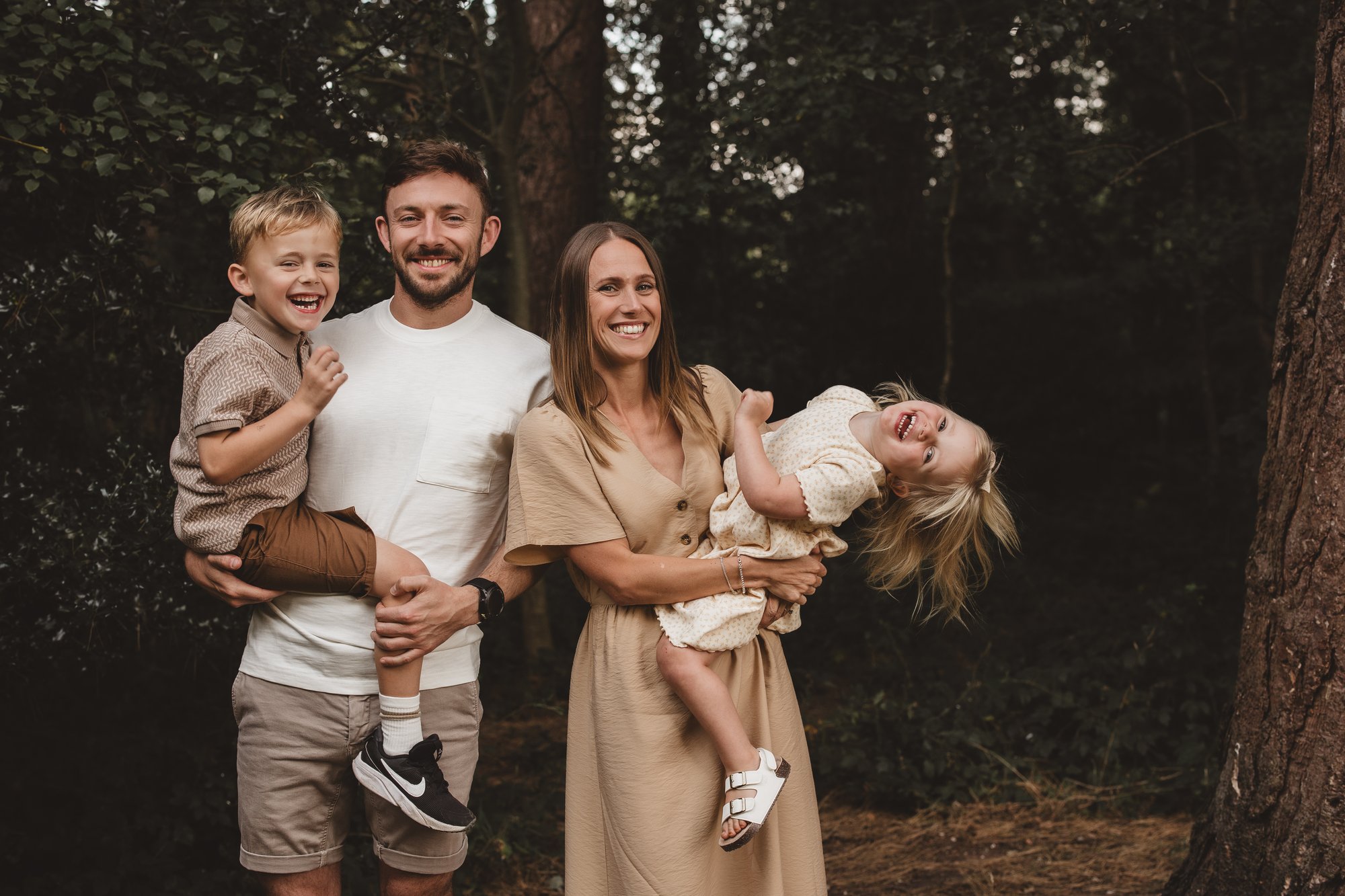 Family of four laughing together in woodland, parents holding their two young children