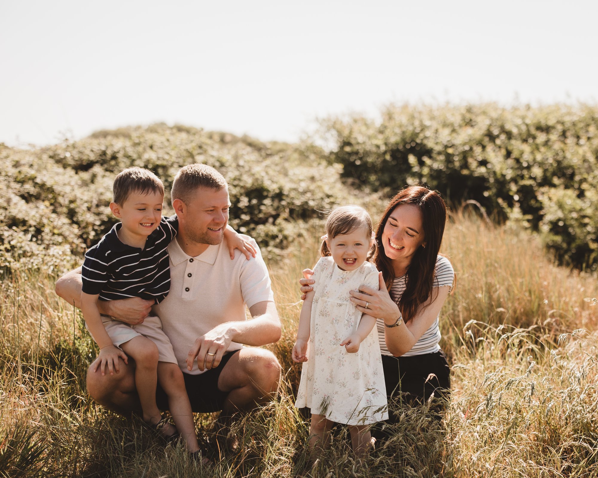 Family of four sitting in sunlit summer grass, parents with their young son and daughter