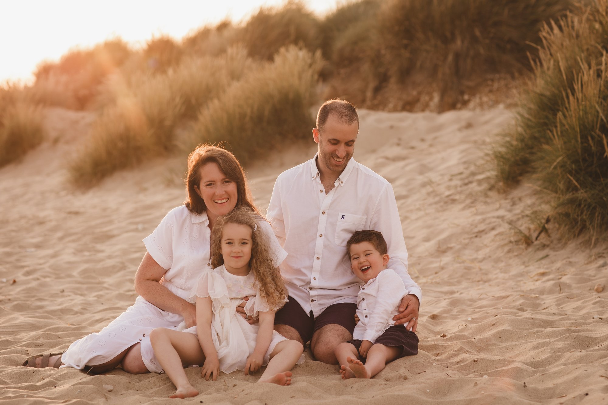 Family of four sitting on a sandy beach at golden hour with dunes behind