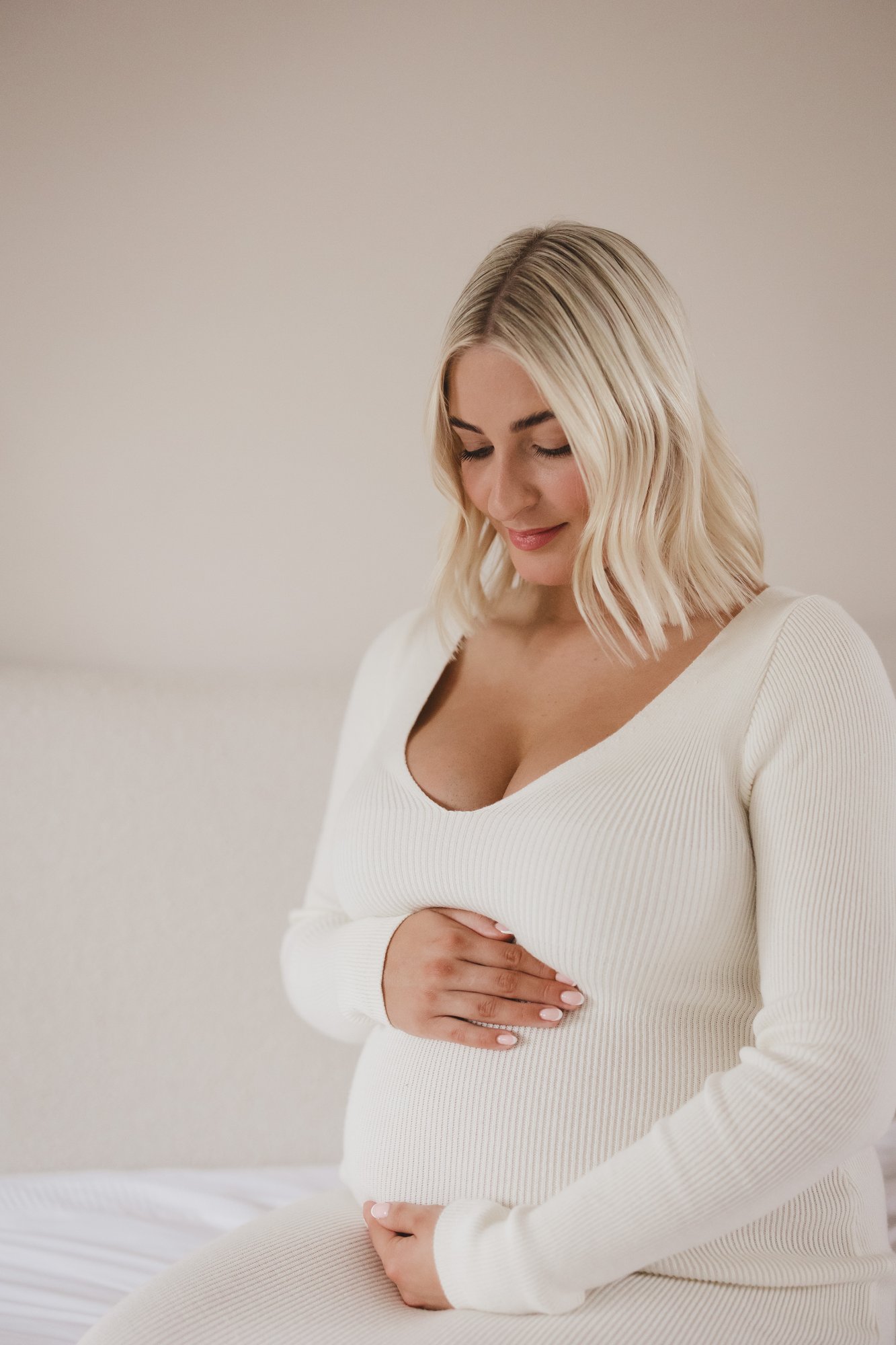 Expectant mother in a cream ribbed dress sitting softly with both hands resting on her bump