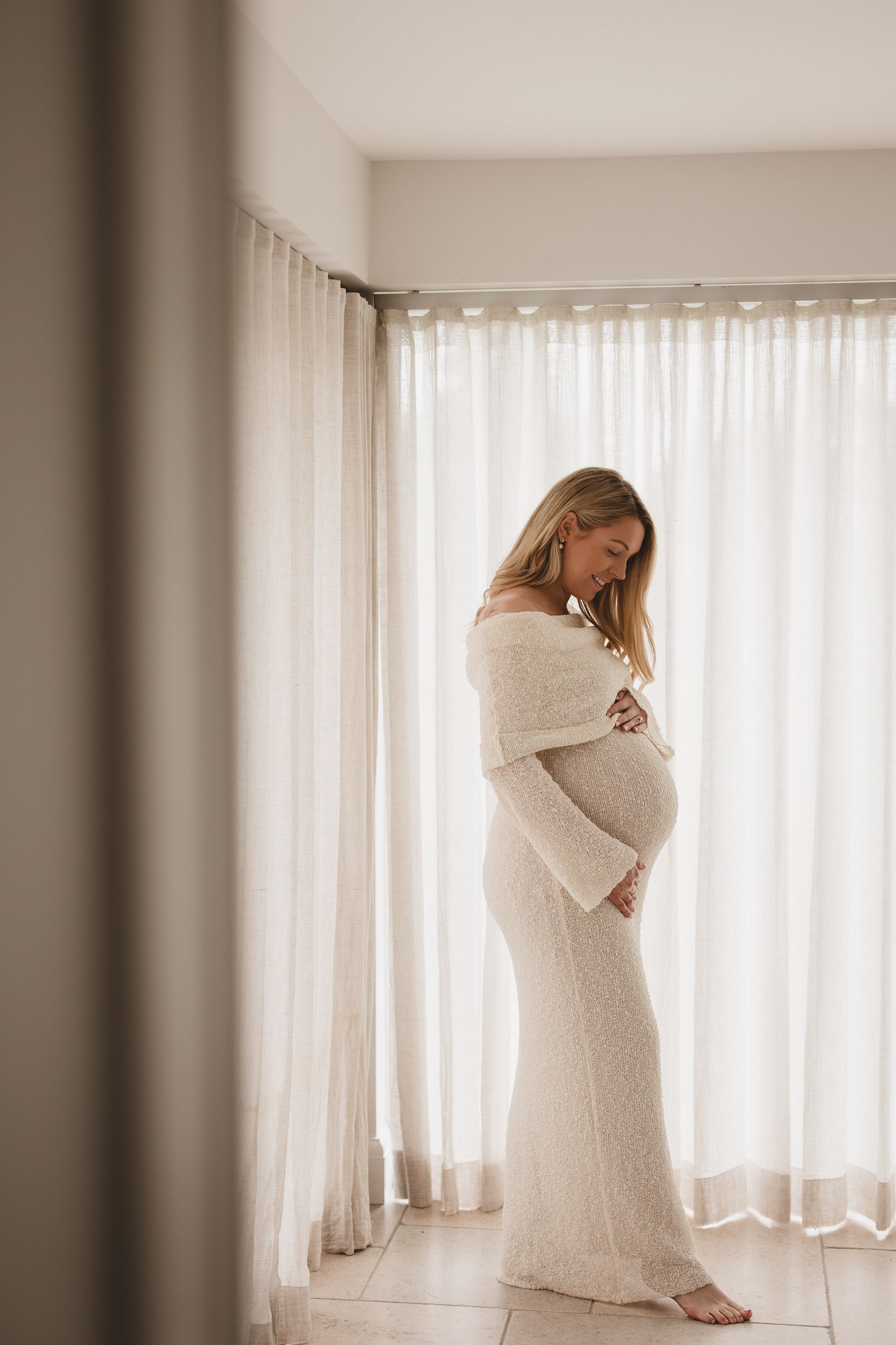 Expectant mother in a cream knit dress standing gently in window light
