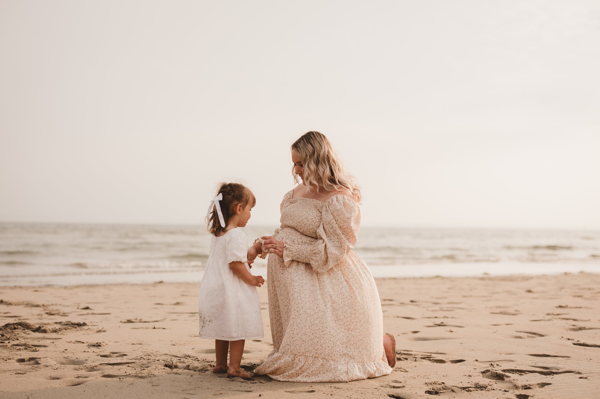 Expectant mother kneeling on the sand with her toddler daughter at sunset