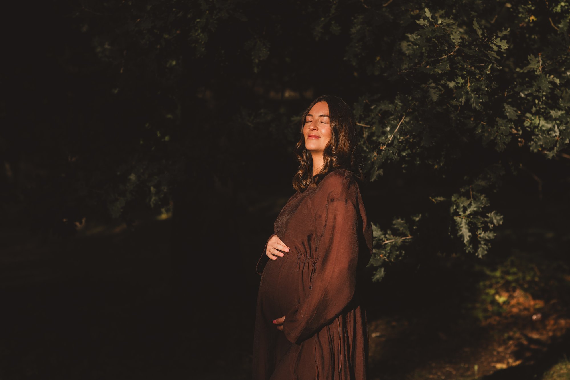 Expectant mother in a flowing brown dress standing peacefully in woodland with dappled light