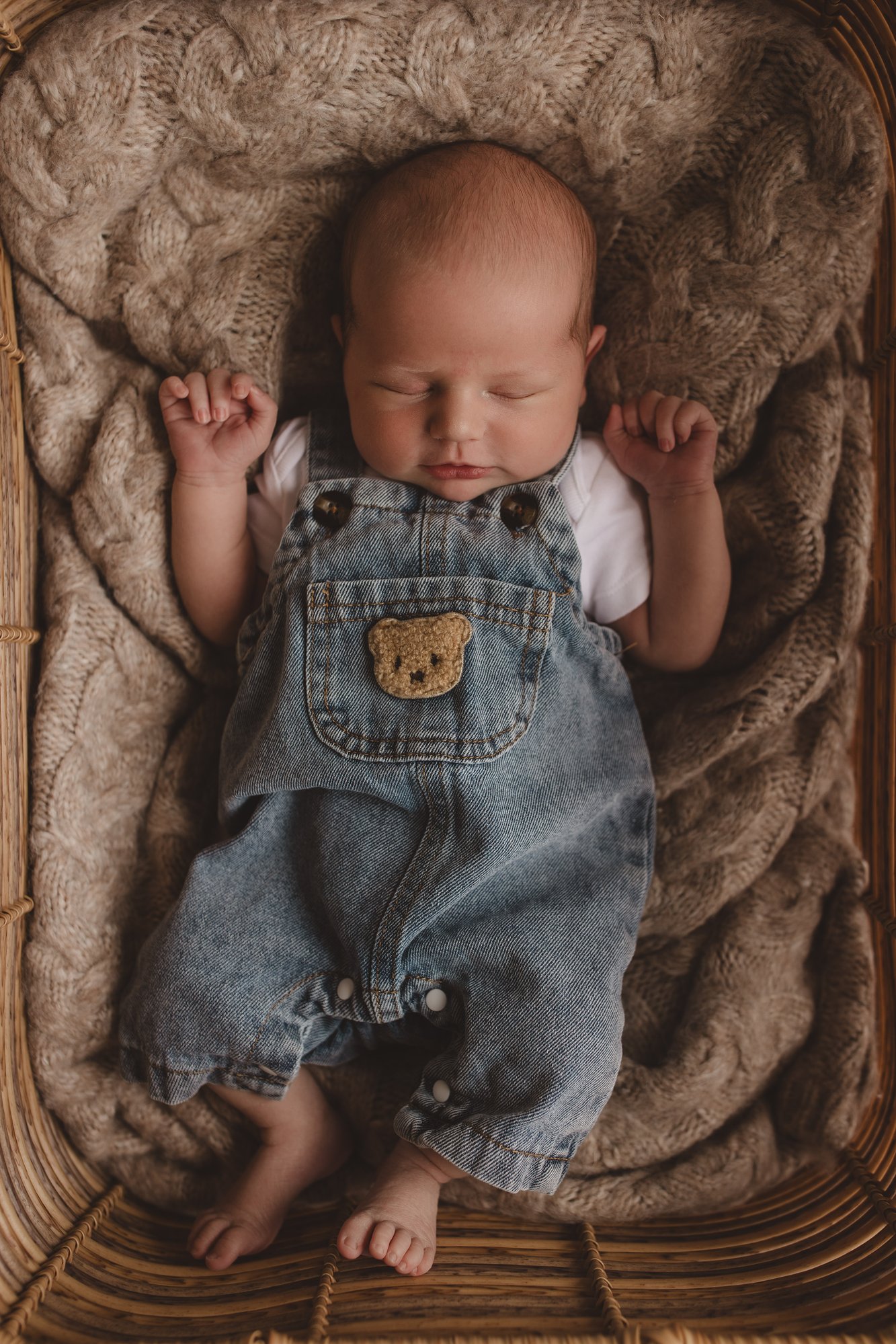 Newborn baby sleeping in a wicker basket wearing denim teddy-bear dungarees, overhead studio shot