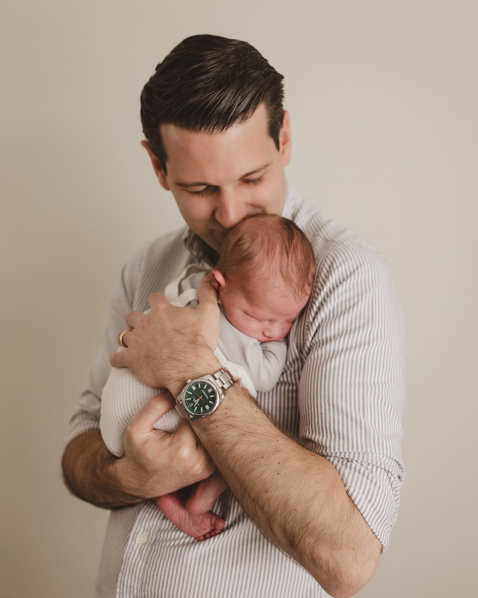 New dad in striped shirt cradling and gently kissing his newborn baby's head