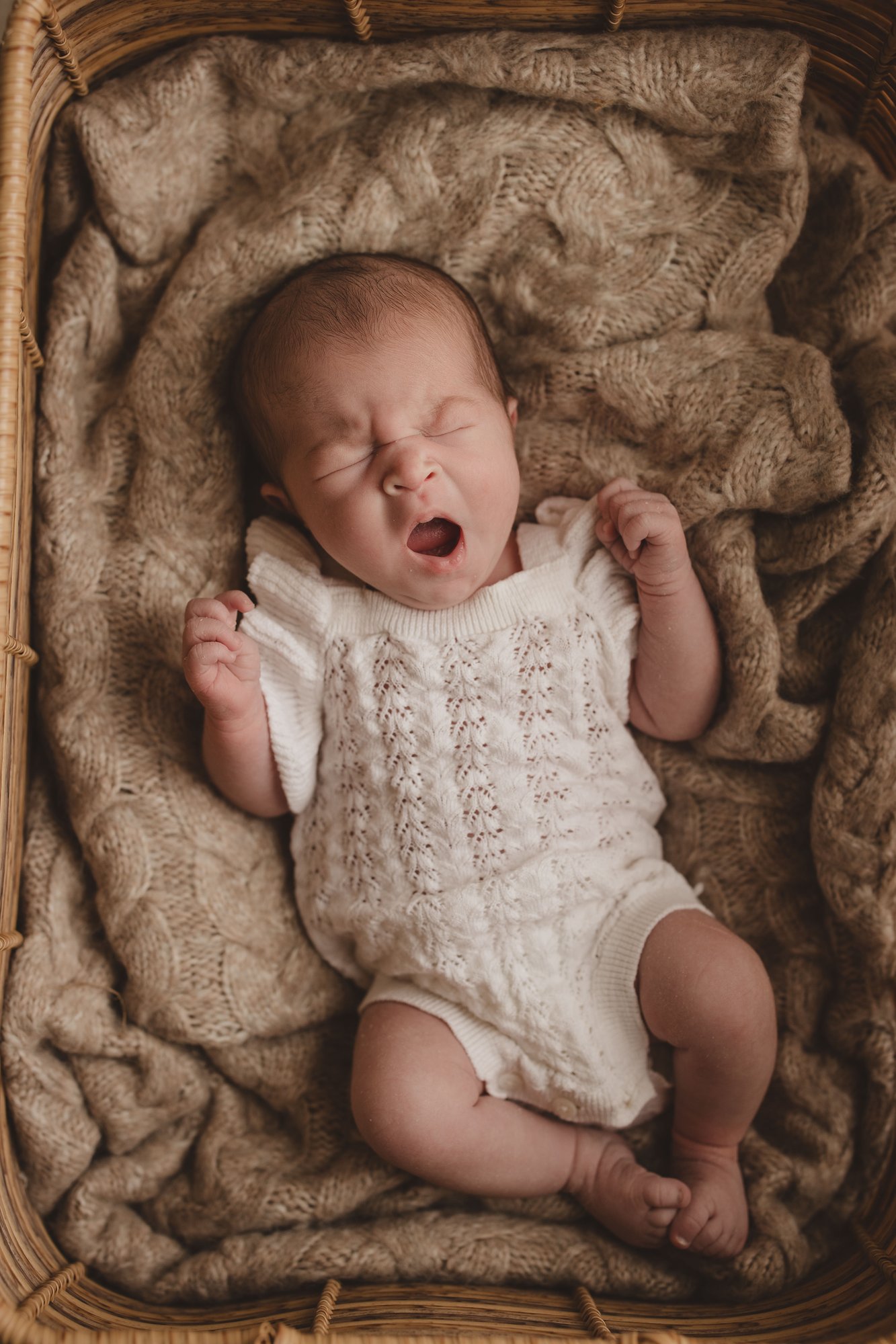Newborn baby mid-yawn in a wicker basket wearing a cream pointelle knit romper