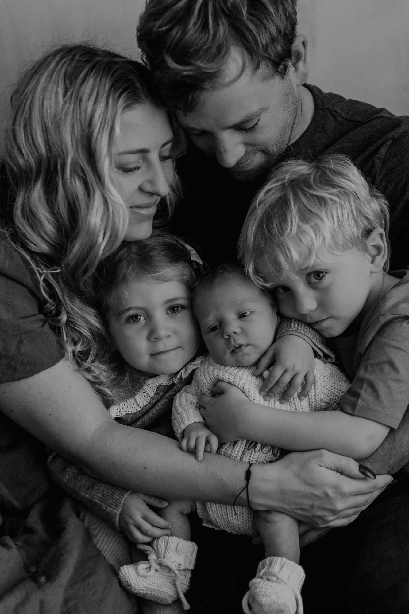 Black and white family portrait of parents with their three young children including the newborn, all gently cuddled together
