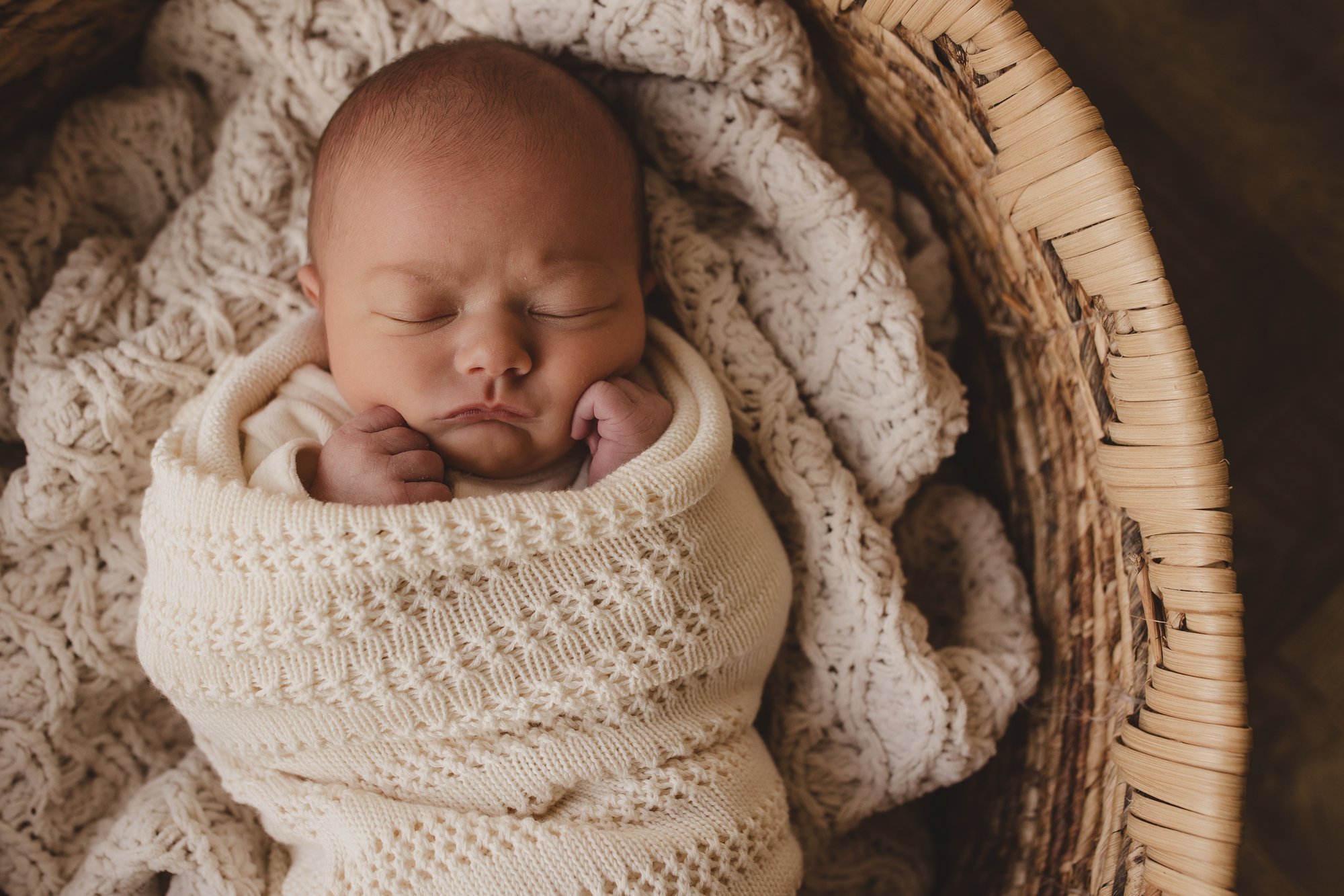 Newborn baby swaddled in a cream knit wrap, peacefully asleep in a woven basket