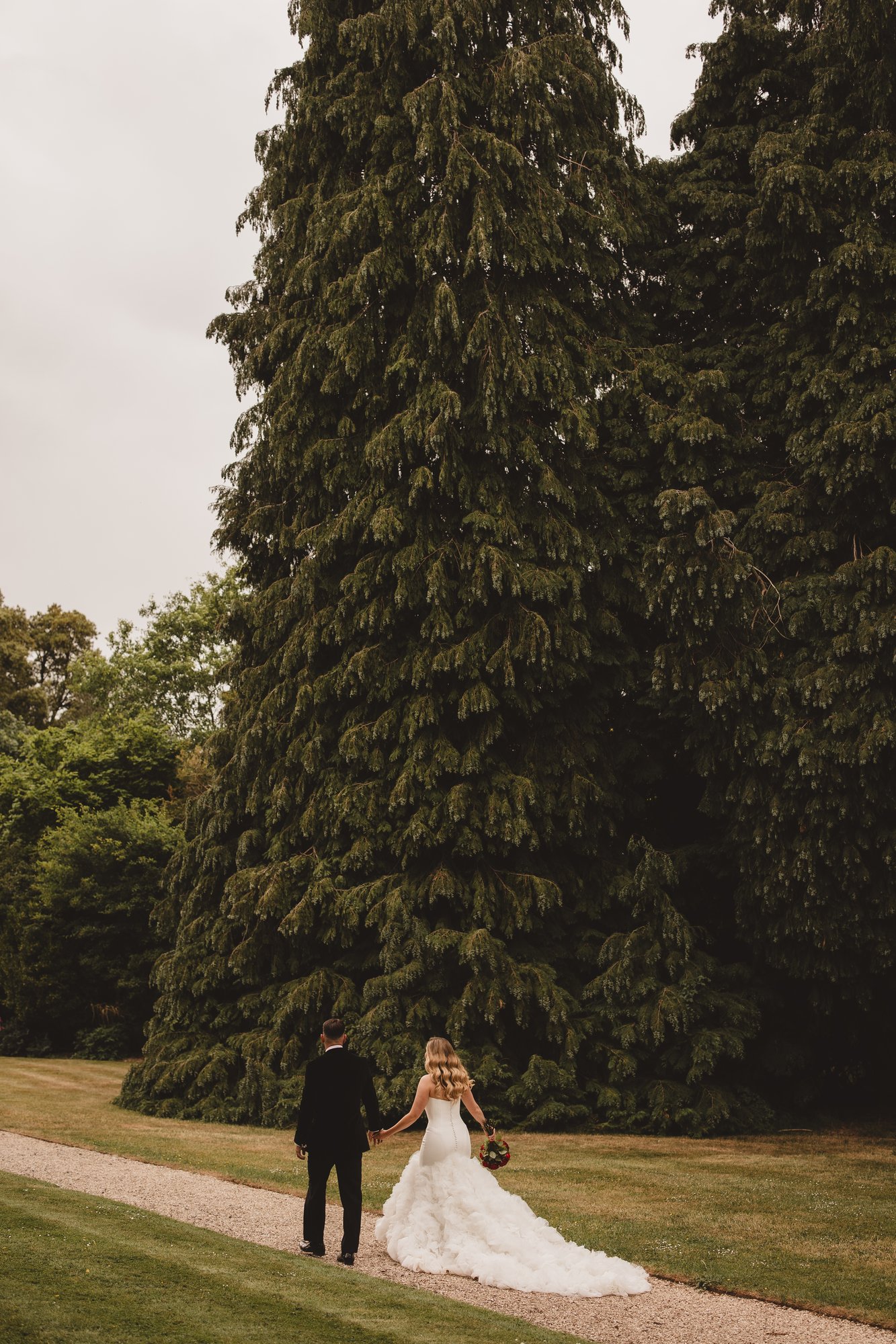 Bride and groom walking past an enormous evergreen tree in formal grounds